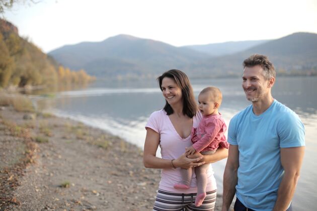 户外一个快乐的家庭 带着一个小孩站在阳光下群山环绕的湖边父母休闲旅行