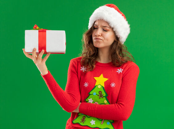 怀疑穿着红色圣诞毛衣 戴着圣诞帽的年轻女子拿着礼物 带着怀疑的表情站在绿色的背景下看着它圣诞老人圣诞节年轻人