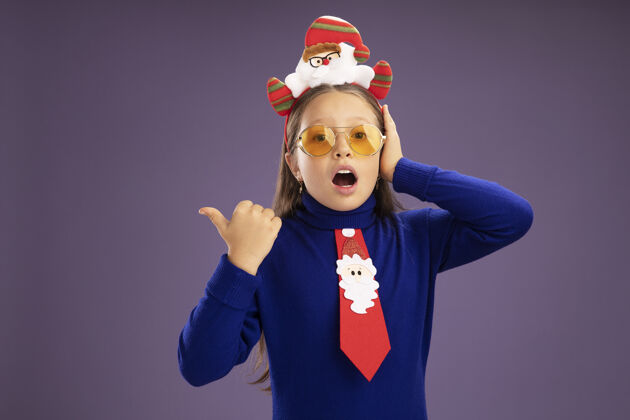 高领毛衣小女孩穿着蓝色高领毛衣 打着红色领带 头上戴着有趣的圣诞戒指 惊讶地看着相机 用大拇指指着站在紫色背景上的一边头领带搞笑