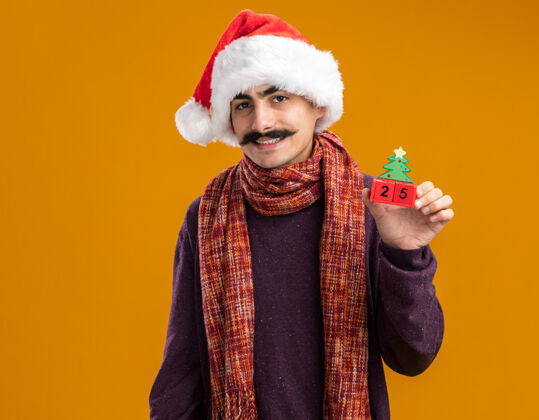 站着留着胡子的男人戴着圣诞老人帽 脖子上围着暖和的围巾 手里拿着和25岁约会的玩具立方体 站在橙色的背景下 面带微笑地看着相机温暖立方体帽子