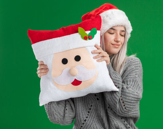 帽子快乐的金发女郎 穿着冬季毛衣 戴着圣诞帽 抱着圣诞枕头 闭着眼睛微笑着站在绿色的背景下金发微笑举行