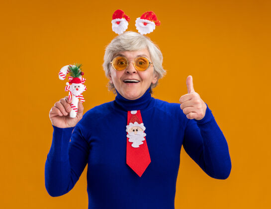 糖果戴着太阳眼镜 戴着圣诞老人头带和圣诞老人领带的老妇人 拿着糖果手杖 竖起大拇指 在橙色背景上孤立地复制空间领带橙色头带