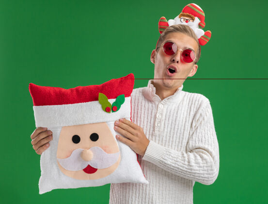 帅气给人印象深刻的年轻帅哥戴着圣诞老人头带戴着眼镜抱着圣诞老人的枕头隔离在绿色的墙上印象深刻圣诞老人年轻