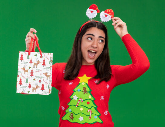 头带戴着圣诞老人头带的快乐的年轻白人女孩拿着纸礼品袋 看着绿色背景上与世隔绝的一面 还有复制空间快乐礼物持有