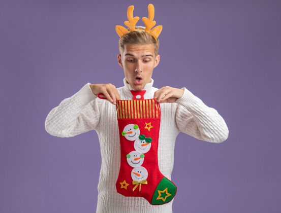 驯鹿给人印象深刻的年轻帅哥戴着驯鹿鹿角头带 打着圣诞老人的领带 手里拿着圣诞长袜 看着里面孤立的紫色墙壁头带圣诞老人穿着