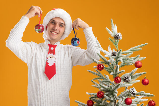 球微笑着的年轻帅哥戴着圣诞帽打着圣诞老人的领带站在装饰好的圣诞树旁拿着圣诞球饰品站在头旁看着隔离在橙色背景上的相机圣诞树抱着帽子