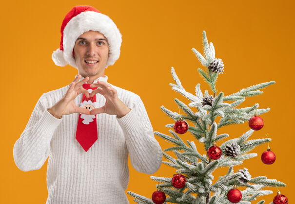 树微笑着的年轻帅哥戴着圣诞帽 打着圣诞老人的领带站在装饰好的圣诞树旁 看着摄像机做着孤立在橙色背景上的心形标志站着小伙子相机