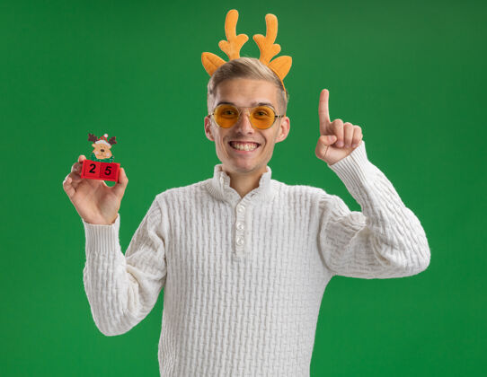 日期快乐的年轻帅哥戴着驯鹿鹿角头带戴着眼镜拿着圣诞树玩具和日期指着隔离在绿色墙上穿着鹿角头带