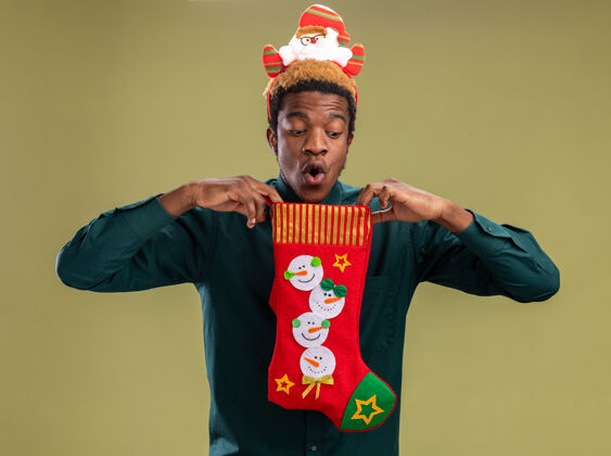 里面站在绿色的背景下 一个非洲裔美国人拿着圣诞长袜 戴着滑稽的圣诞帽 打着红领带 好奇地看着里面搞笑领带站着