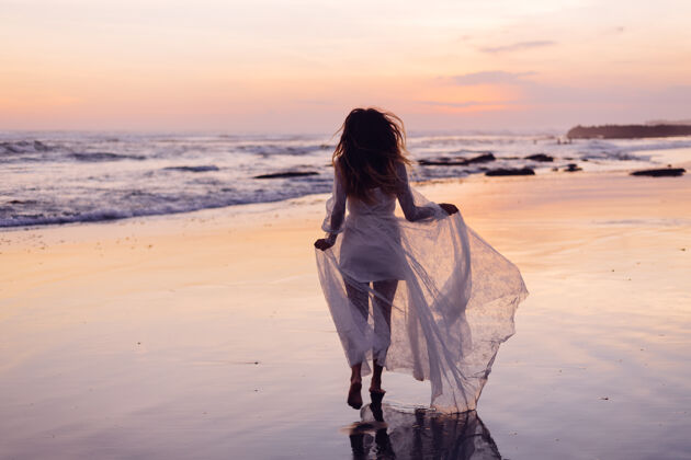 乐趣美丽的白种女人独自一人穿着白色的裙子在紫色的夕阳边远眺优雅户外海浪
