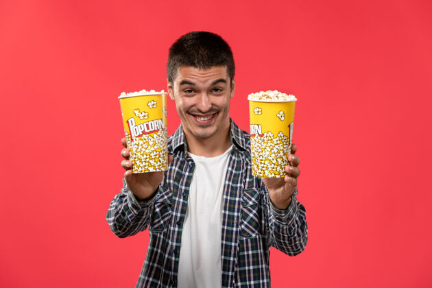 成人正面图年轻男子手持爆米花包 微笑着在淡红墙电影院看电影包装爆米花剧院