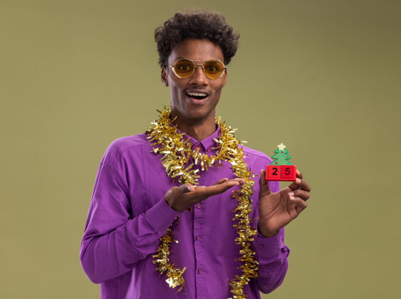 举行快乐的非洲裔美国年轻人戴着一副眼镜 脖子上戴着金属丝花环 拿着并指着圣诞树玩具和日期 看着橄榄绿背景上孤立的相机圣诞节日期指着