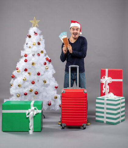 展示带着红色手提箱的快乐男人在灰色的墙上展示他的旅行票帽子圣诞盒子