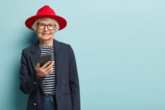 个人老年人和科技概念老年妇女始终保持联系 通过应用程序聊天 发送信息 戴红色头饰 条纹套头衫和正式夹克 隔着蓝色的墙短信时尚浏览