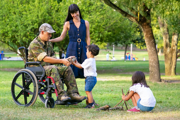 活动两个孩子在妈妈和轮椅上的残疾军人爸爸附近安排户外篝火的木柴男孩向父亲展示木柴残疾退伍军人或家庭户外概念轮椅携带军队