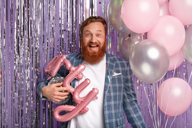 大笑乐观的红发男人 浓密的胡须和时髦的发型 拿着气球和迪斯科舞会 和客人一起狂欢 穿着时髦的衣服 隔着紫色的墙壁生日派对来了胡须狂欢红发