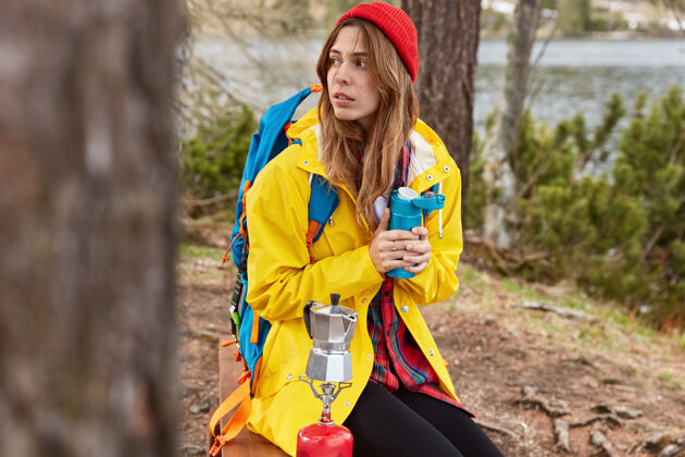 水年轻沉思的女性背着背包坐在河边或湖边的小树林里 用热水瓶的热饮取暖 在野营的炉子上煮咖啡健康饮料举行