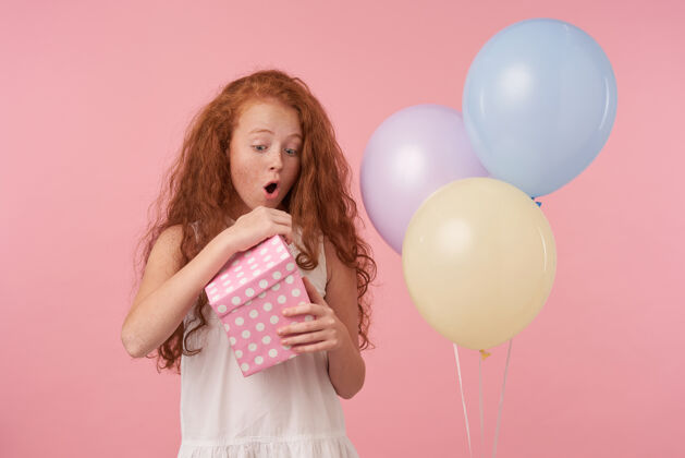 兴奋可爱的红发女孩穿着优雅的礼服庆祝节日的摄影棚照片 打开礼盒兴奋的脸 摆出粉红色背景和彩色气球盒子孩子表情