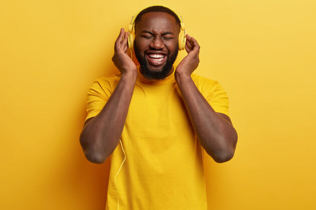 听喜出望外的美国黑人男子的单色镜头 戴着新耳机享受完美的响亮声音 穿着黄色t恤 有空 用音乐娱乐自己快乐的表情唱歌节奏丰满