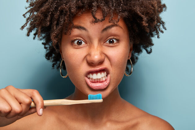 非洲健康女孩的画像刷牙 拿着木刷子 有早晨的例行公事 卷发 在室内蓝色墙壁上摆姿势 展示裸露的肩膀早起人 种族和卫生观念室内头像高兴
