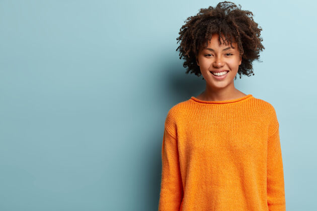 肖像可爱的积极的非洲裔美国妇女的照片 卷发 穿着橙色套头衫 精神饱满 站在蓝色的墙上 为您的宣传内容提供免费空间好的情感概念高兴黑暗大笑
