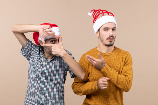 手前视图两个戴着圣诞帽的家伙一个用手做摄像机标志 另一个用米色孤立的背景展示他两个伙计们圣诞老人前面