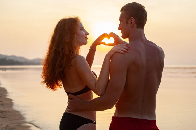 夏天年轻性感浪漫的情侣在夏天的沙滩上快乐地在一起 穿着泳衣在日落时露出心形标志约会浪漫海洋