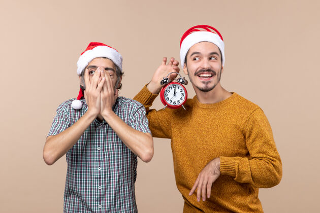 手前视图两个圣诞老人一个用手捂住一只眼睛 另一个拿着一个闹钟 背景是米色的两个男人两个圣诞男人