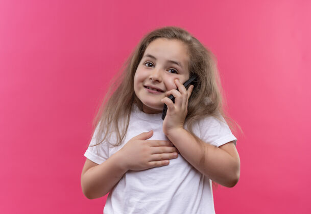 穿着微笑的小女孩穿着白色t恤在孤立的粉红色背景下讲电话微笑小电话
