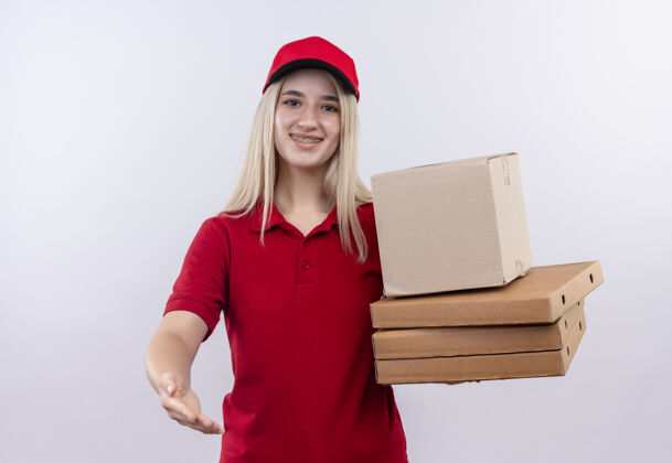 年轻微笑着送货的年轻女孩穿着红色t恤 戴着帽子 戴着牙套 手里拿着披萨盒 在孤立的白色背景下 向镜头伸出手来手盒子红色