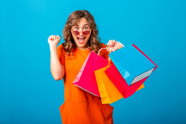 持有图片中的兴奋迷人的微笑时尚女性购物狂穿着橙色的新潮连衣裙拿着购物袋在蓝色的工作室背景隔离销售开朗女性