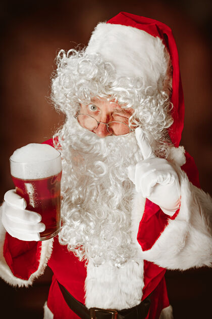 礼物穿着圣诞老人服装的人的肖像-一个豪华的白胡子 圣诞老人的帽子和红色服装在红色工作室背景与啤酒欢乐的肖像场合