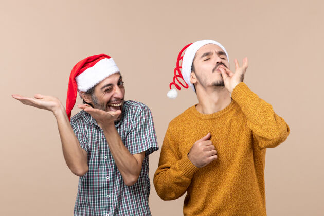 男人前视图两个带着圣诞帽的微笑的家伙 一个在展示什么 另一个在米色孤立背景上制作厨师亲吻标志帽子两个微笑的家伙成人