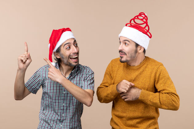 前面前视图两个带着圣诞帽的微笑的家伙 一个在米色孤立的背景上向另一个展示东西帽子两个微笑的家伙米色