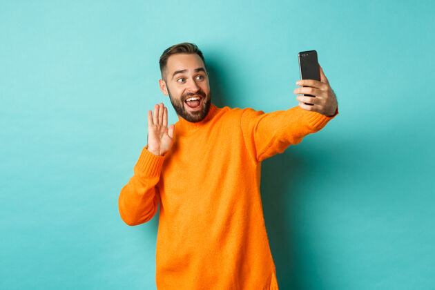 智能手机快乐的年轻人视频通话 用手机上网聊天 和智能手机摄像头打招呼 友好地挥手 站在浅蓝绿色的墙上成人男性室内