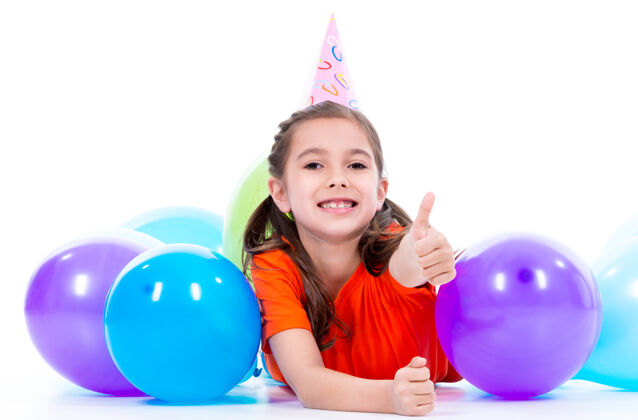 彩色穿着橙色t恤的快乐微笑女孩躺在地板上 手里拿着五颜六色的气球 竖起大拇指——被隔离在白色的地板上手势休闲气球