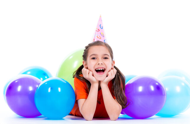 积极穿着橙色t恤的快乐微笑女孩躺在地板上 带着五颜六色的气球-孤立在白色的地板上小五颜六色漂亮