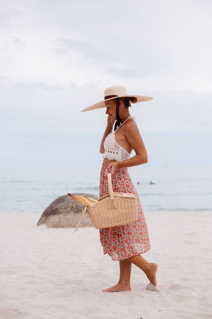 肖像沙滩上的浪漫女人穿着裙子 针织上衣 戴着草帽 抱着面包篮子 生态生活散步热带放松
