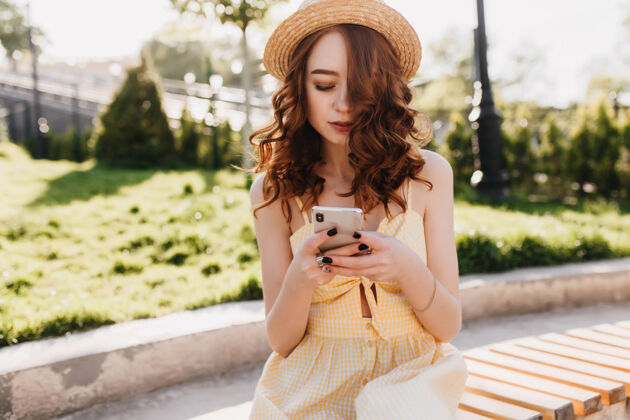 姜美丽的公园里 一位戴着时髦帽子的年轻女士坐在那里发短信一张姜黄色头发的时髦女孩在长凳上等着别人的户外照片成人可爱波浪
