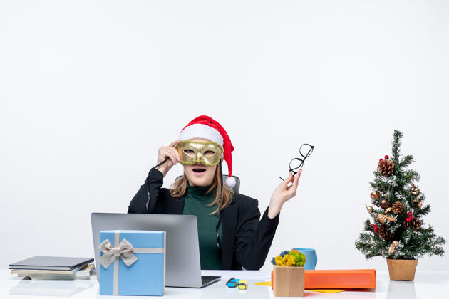 笔记本在白色背景的办公室里 一位戴着圣诞老人帽 戴着眼镜 戴着面具的年轻女士坐在一张桌子旁 桌上放着圣诞树和一份礼物圣诞节圣诞树圣诞老人