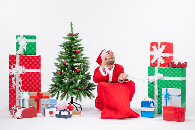 坐着专注的年轻人打扮成圣诞老人与礼物和装饰圣诞树坐在地上 看着上面的白色背景看季节圣诞老人