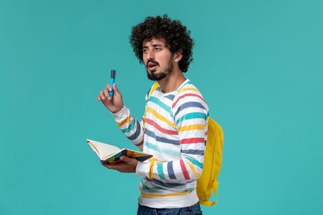 学校穿着条纹衬衫 背着黄色背包 拿着毛毡笔和蓝色墙上的抄写本的男学生的正视图学生大学书籍