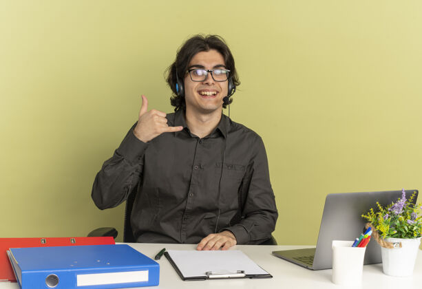 标志戴着耳机 戴着眼镜 面带微笑的年轻上班族坐在办公桌旁 手里拿着办公工具 手里拿着笔记本电脑手势和手机手势年轻工人工具