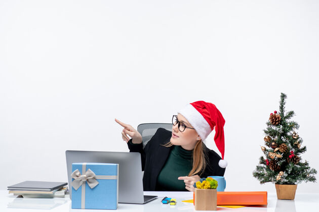圣诞树金发碧眼的年轻女子 戴着圣诞老人的帽子 坐在一张桌子旁 桌子上放着圣诞树和礼物 在白色背景上指着上方笔记本金发坐着