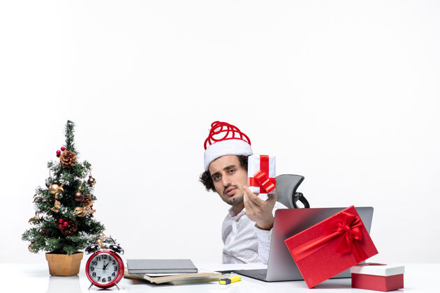 帽子新年心情与悲伤不满的年轻商人圣诞老人的帽子坐在办公室里 并提出他的礼物白色背景提高办公室悲伤
