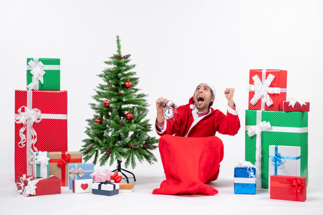 圣诞老人紧张的圣诞老人坐在地上 在礼物和装饰圣诞树旁的白色背景上展示时钟圣诞地紧张