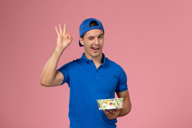 粉色正面图身穿蓝色制服披肩的年轻男性信使手持圆形的投递碗在浅粉色的墙上眨眼快递员视图人