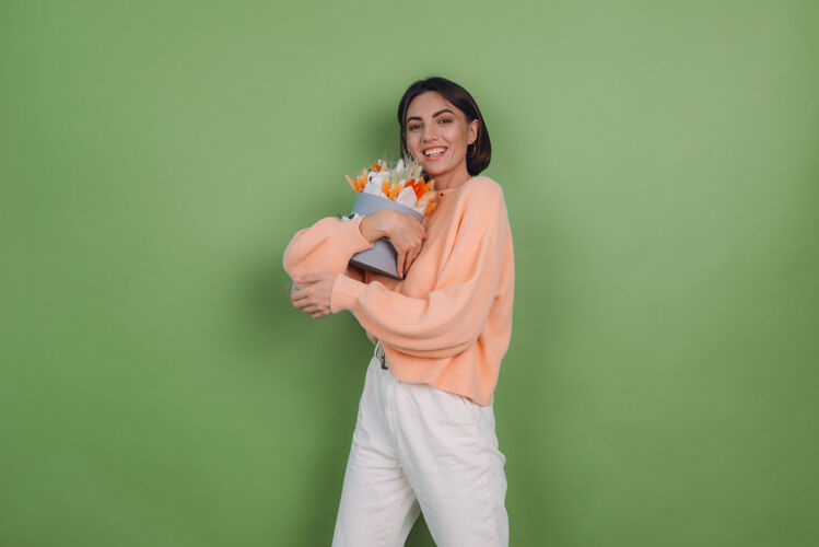 女性年轻女子穿着休闲桃色毛衣隔离在绿橄榄墙上手持橙白色花盒组成的棉花 吉普赛拉小麦和拉古鲁斯作为礼物开心惊喜静物帽盒内部