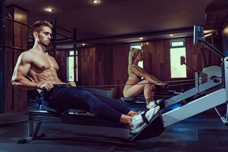 背部肌肉发达的健美运动员在运动服训练后坐在锻炼机上在黑暗的氛围中 在健身房里拥有完美健身身体的男人的侧视图健美 健身的概念运动员运动身体