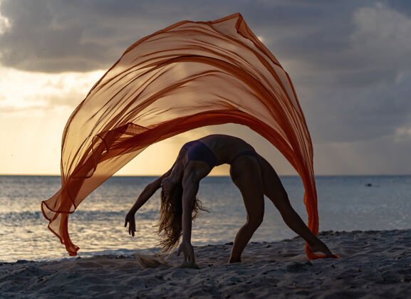 自由柔韧的女杂技演员的剪影在戏剧性的日落中用丝绸做戏法 海景上有暴风雨的云水织物杂技
